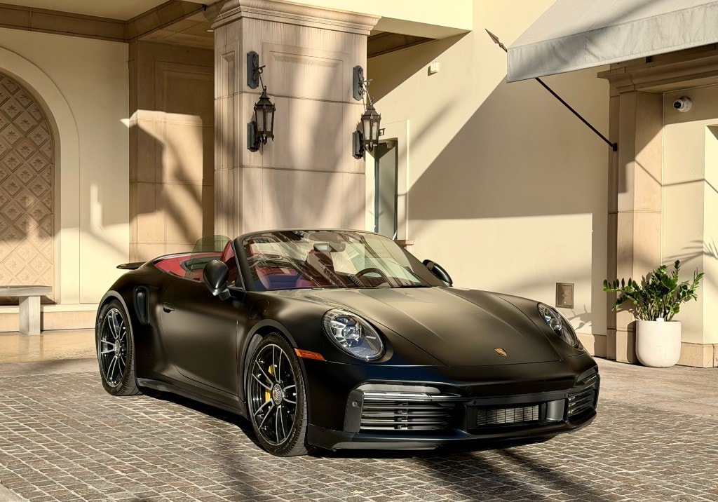 Porsche 911 Turbo S Cabriolet — matte black front three-quarter at a luxury hotel entrance